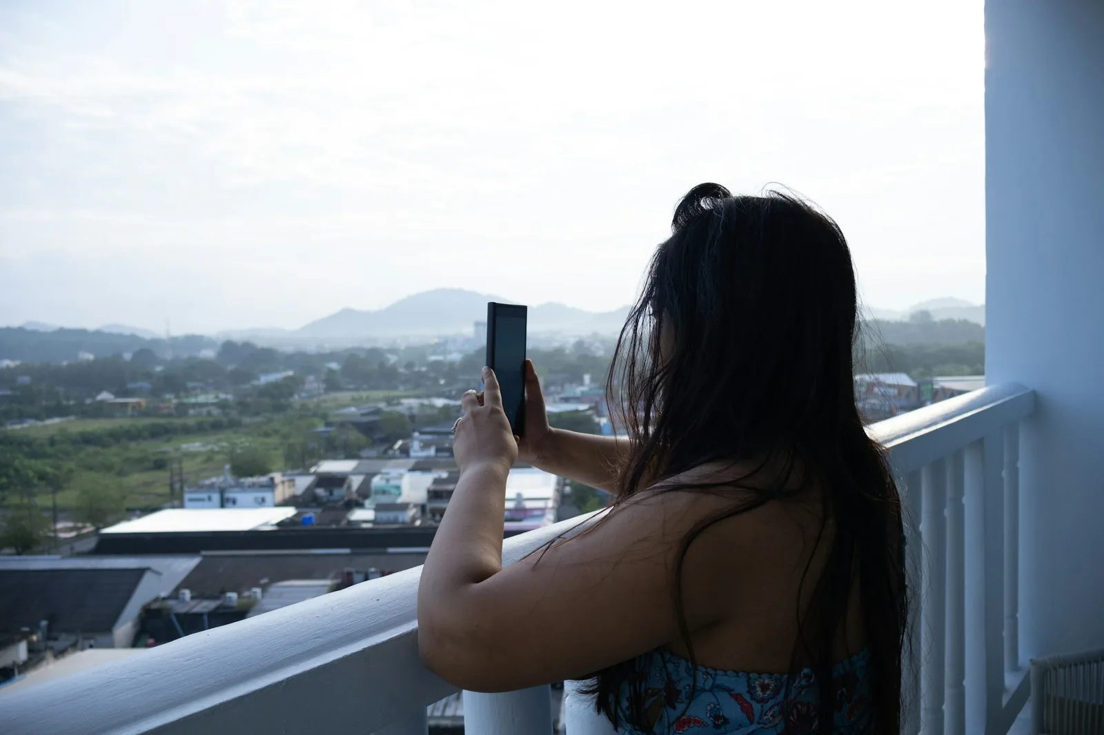 Mulher de costas numa varanda alta segura o celular diante da cidade e das montanhas.