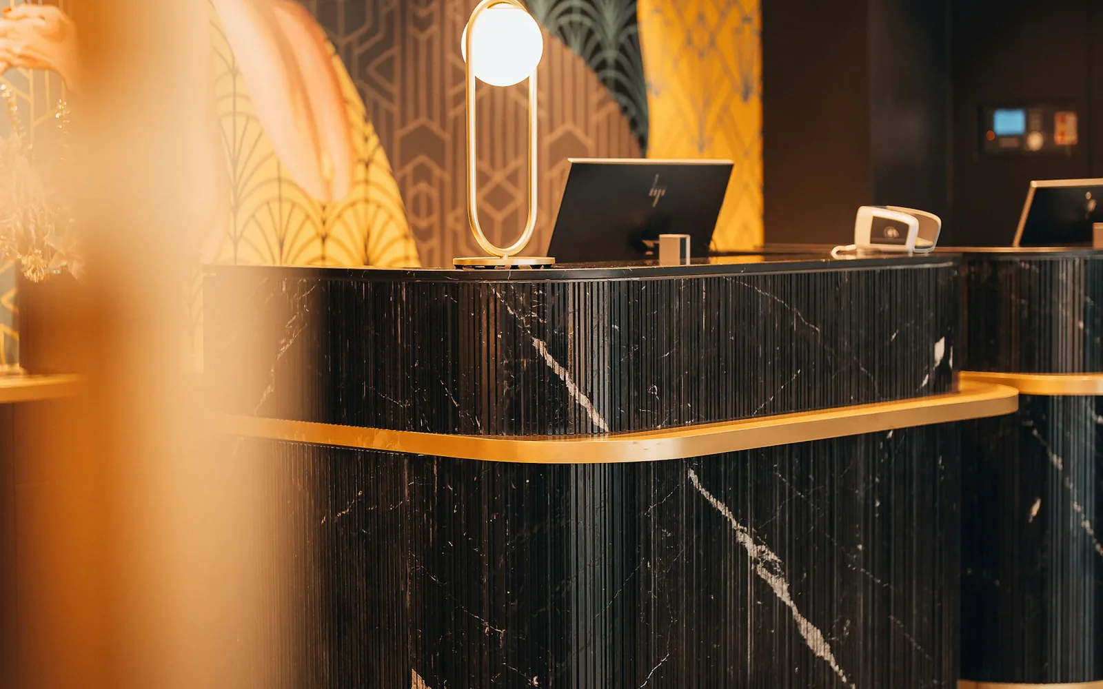 Balcão de recepção de hotel em mármore escuro com acabamento dourado e luminária acesa.